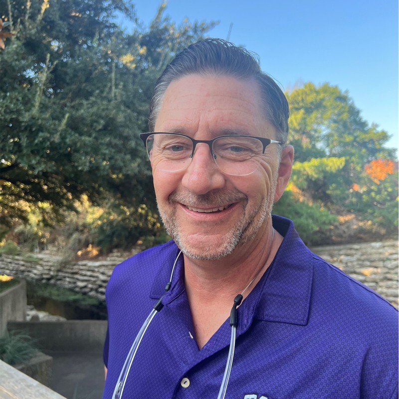 Smiling man wearing glasses and purple shirt outdoors with greenery.