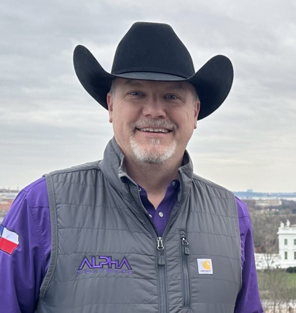 Man in cowboy hat wearing Alpha Polymers vest smiling outdoors.