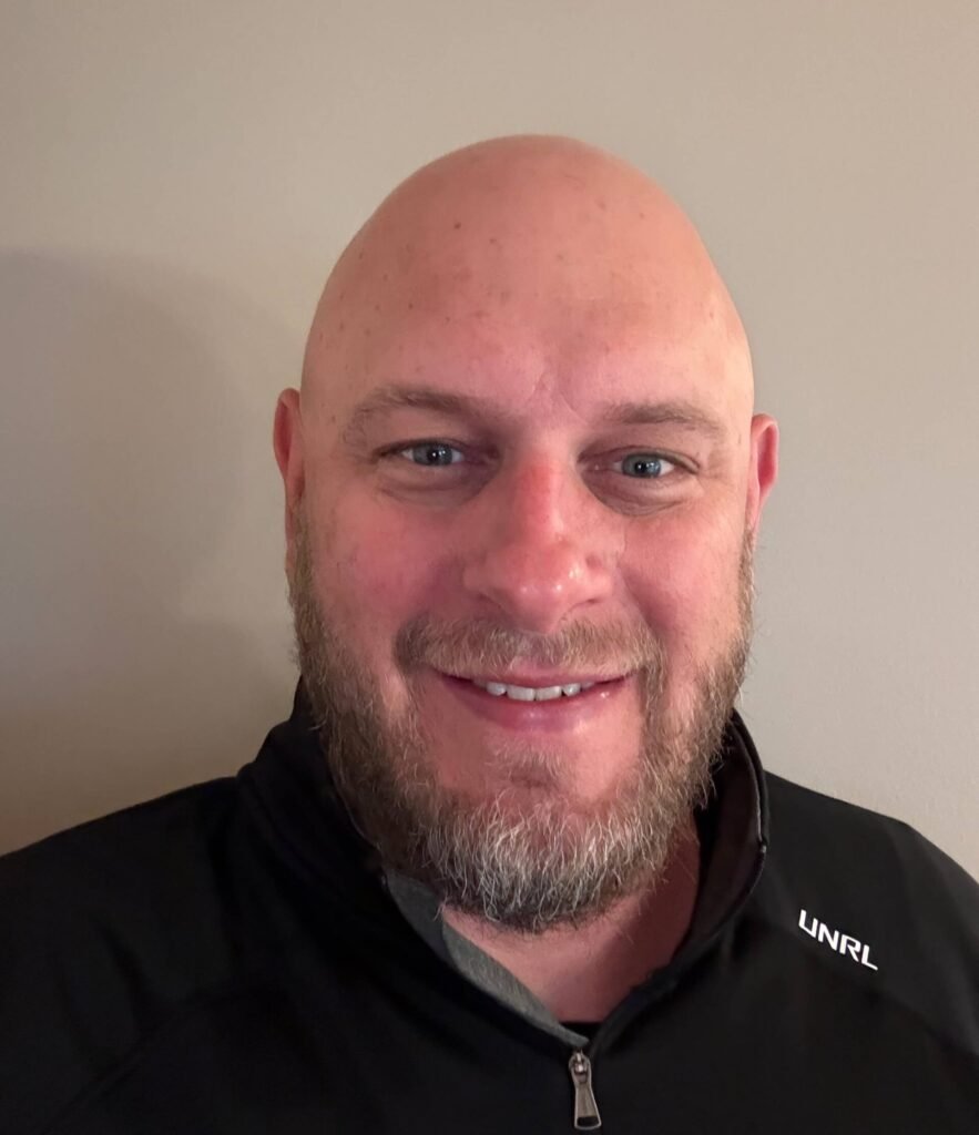Smiling bald man with beard wearing black UNRL jacket indoors.