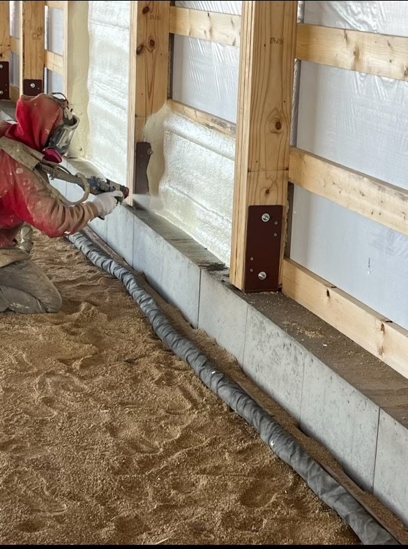 Worker in protective gear applying spray foam insulation along wooden wall base.