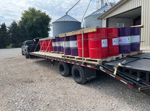 Flatbed truck transporting red and purple chemical barrels outside farm buildings.